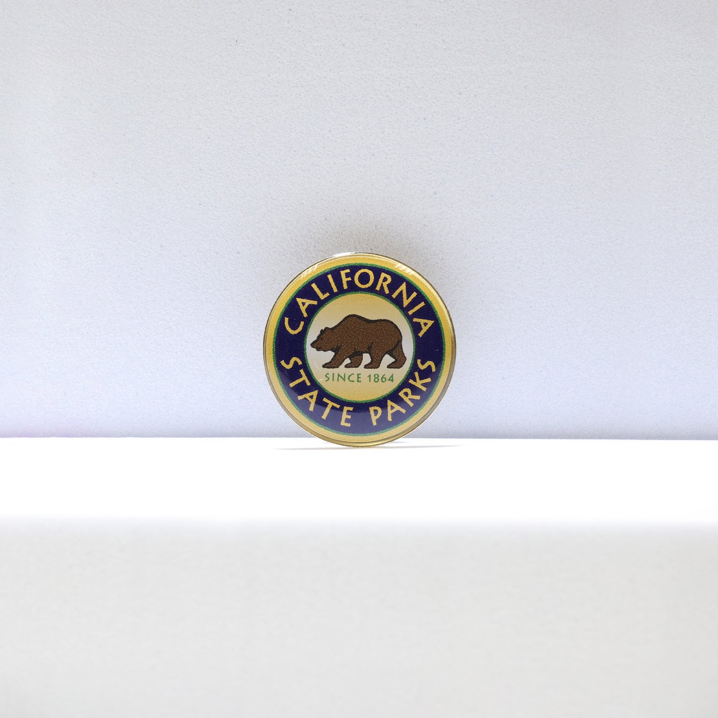 a round green, blue, and yellow pin, with a brown grizzly bear in the center atop the words Since 1864, and the words California State Parks circling around the rim of the pin