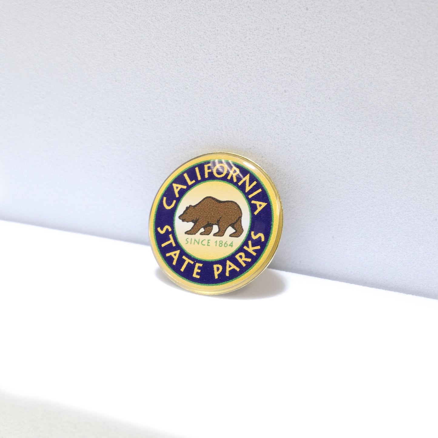a round green, blue, and yellow pin shown at an angle, with a brown grizzly bear in the center atop the words Since 1864, and the words California State Parks circling around the rim of the pin
