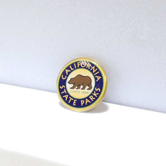 a round green, blue, and yellow pin shown at an angle, with a brown grizzly bear in the center atop the words Since 1864, and the words California State Parks circling around the rim of the pin